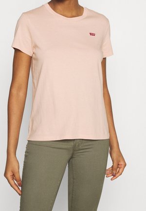 Kvinde iført en lyserød T-shirt med korte ærmer og et lille rødt Levi's-logo på brystet samt olivengrønne bukser.