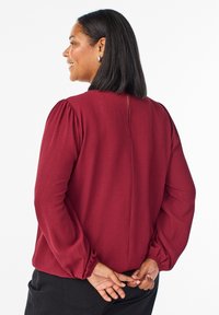 Kobieta o ciemnych włosach sięgających do ramion, nosząca srebrne kolczyki w kształcie koła i bordową bluzkę z długim rękawem, z rękami splecionymi za plecami.