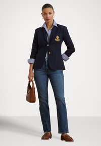 Blazer azul-marinho com botões dourados e logótipo bordado, combinado com camisa às riscas azul, jeans azuis e mocassins castanhos, segurando uma mala castanha.