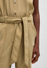 Beige jumpsuit with a tied waist belt, featuring a button-up front and subtle texture. The fabric appears soft and lightweight.