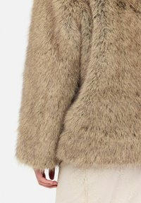 Cappotto in finta pelliccia di un leggero beige con una superficie morbida e testurizzata. Caratterizzato da maniche larghe e una vestibilità cropped, accentuato da una mano posata su una gonna chiara.