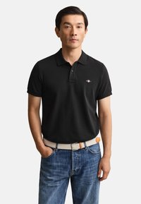 Man standing with hands in pockets wearing black polo shirt, blue jeans, and beige woven belt against plain background.