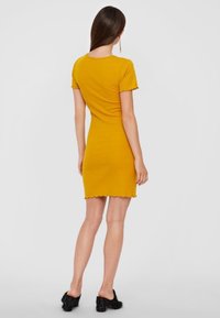 Gul figursydd klänning med korta ärmar, scallopad nederkant och ribbad textur, bärs med svarta texturerade skor. Bakvy.