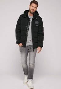 Giacca a vento nera con cappuccio, dotata di chiusura con zip, dettagli del logo sulle maniche, indossata su una maglietta grigia a maniche lunghe e jeans grigio chiaro. Sneakers bianche.