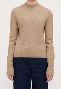 Maglione beige con texture a coste, colletto alto e maniche lunghe. Presenta una silhouette aderente e polsini allungati. Indossato sopra jeans scuri.