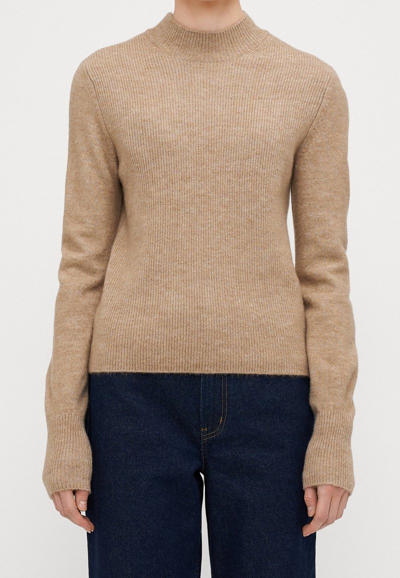 Maglione beige con texture a coste, colletto alto e maniche lunghe. Presenta una silhouette aderente e polsini allungati. Indossato sopra jeans scuri.