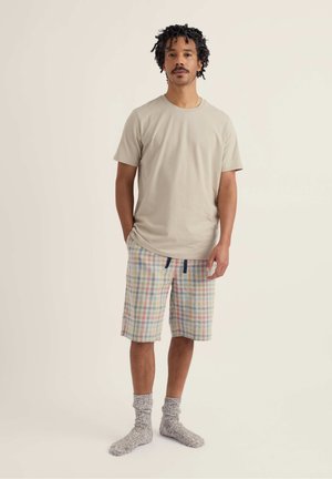 Homme aux cheveux courts et bouclés portant un t-shirt beige, un short à carreaux avec cordon de serrage et des chaussettes grises, debout devant un fond uni.