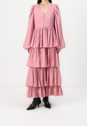 Vrouw draagt een lange roze jurk met laagjes, met opgeblazen lange mouwen en knopen op het lijfje, gecombineerd met zwarte puntige enkellaarzen.