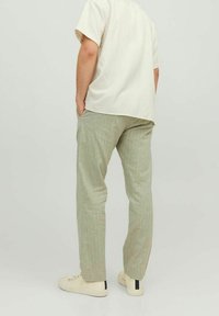 Person står med händerna i fickorna iförd en beige kortärmad skjorta, ljusgröna byxor och off-white sneakers mot en enfärgad bakgrund.