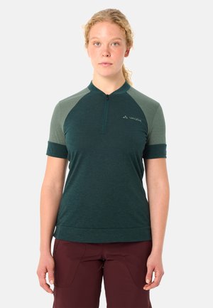 Frau mit lockigem Haar, die ein grünes Kurzarmshirt mit Reißverschluss und bordeauxrote Hosen trägt und vor einem schlichten weißen Hintergrund steht.