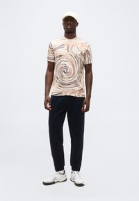 Άνδρας που φοράει μπεζ μπλούζα με σπειροειδές μοτίβο, μαύρο παντελόνι, λευκά παπούτσια EA7 και μπεζ καπέλο, στέκεται σε απλό γκρι φόντο.