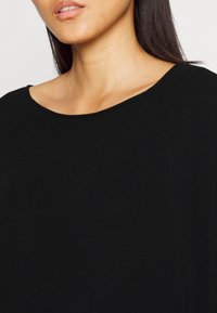 Schwarzes Strickoberteil mit weitem Halsausschnitt, gerippter Textur und glatter Oberfläche, das eine Nahaufnahme des Stoffs und der Designdetails zeigt.