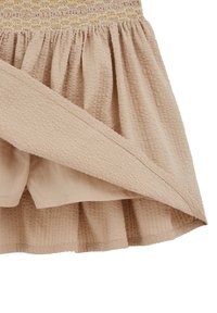 Gaiši bēša svārki ar teksturētu audumu un savāktu vidukli. Ir stingrs augšdaļa ar dzelteniem akcentiem un izplešamu apakšu.