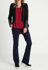 Svart blazer med läderdetaljer, röd blus med knappar, marinblå byxor och svarta sneakers med vita sulor. Släta tyger.