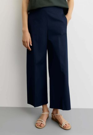 Persona che indossa pantaloni a gamba larga blu navy corti con pince frontali e sandali beige con cinturini intrecciati, in piedi davanti a uno sfondo semplice.