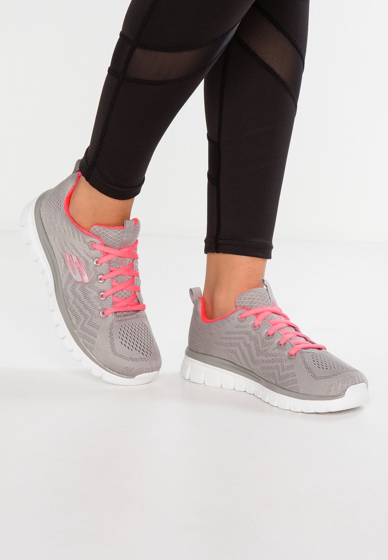 Graue Sportschuhe mit einem strukturierten Strickoberteil und leuchtend rosa Schnürsenkeln, ausgestattet mit einer weißen Gummisohle und einem rutschfesten Design.