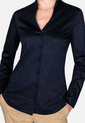 Personne portant une chemise noire à manches longues boutonnée rentrée dans un pantalon beige, les mains dans les poches, sur un fond uni.