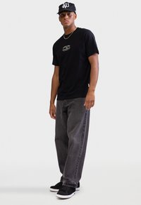 Μαύρο T-shirt με λογότυπο, γκρι τζιν και μαύρα αθλητικά παπούτσια. Το μοντέλο φοράει μαύρο καπέλο με λευκές γράμματα. Απλή, καθημερινή αστική εμφάνιση.