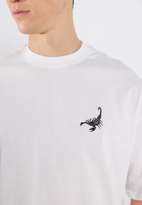 Vit bomullströja med en liten svart skorpiongrafik på vänster bröstområde; rundhalsad design med korta ärmar.