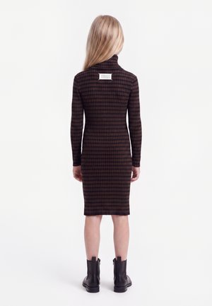 Fille aux longs cheveux blonds portant une robe à manches longues à carreaux marron et noir et des bottines noires, vue de dos.
