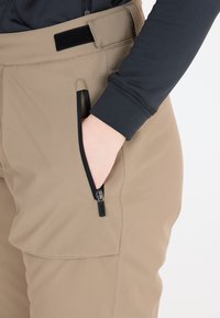 Beige housut, joissa on sileä pinta, sivutasku mustalla vetoketjulla. Musta pitkähiha Polo-paita on osittain näkyvissä.