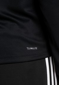 Camiseta atlética negra con una textura suave, con un logo blanco de 'Climalite'. Posicionada cerca de un pantalón negro con tres rayas verticales blancas.