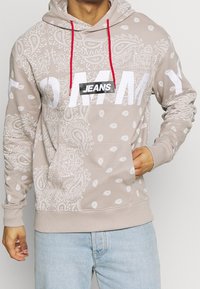 Felpa beige con motivo paisley bianco, lacci rossi e grande testo bianco "TOMMY". Presenta un'etichetta nera "JEANS" sul davanti.