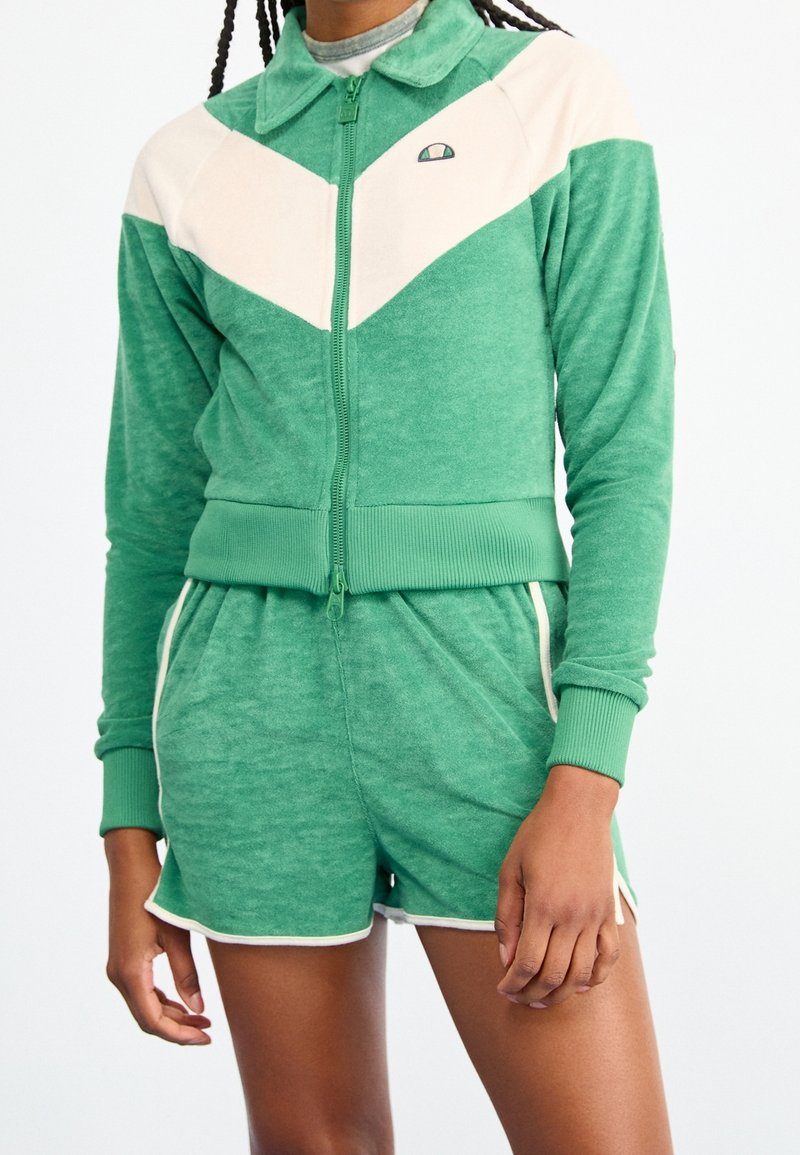 Grön velourträningsdräkt med en zip-up jacka som har krämfärgade chevronränder och ribbade manschetter, tillsammans med matchande shorts, kantade i vitt.