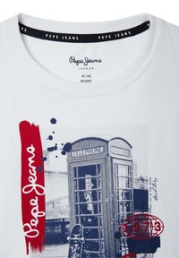 T-shirt de algodón blanco con diseño impreso que presenta un acento rojo, una cabina telefónica de Londres azul y elementos de texto en negro y rojo.
