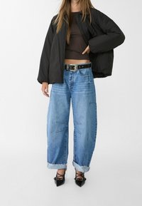 Giacca a vento nera, top corto marrone, jeans in denim azzurro oversize con orli risvoltati e tacchi neri a punta con dettagli in stud.