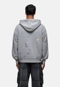 Uomo che indossa una felpa grigia con macchie di vernice multicolore e pantaloni cargo neri, visto di spalle su uno sfondo neutro.
