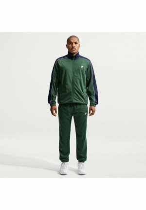 Homme portant un survêtement zippé vert foncé et bleu marine avec des baskets blanches, debout devant un fond clair uni.