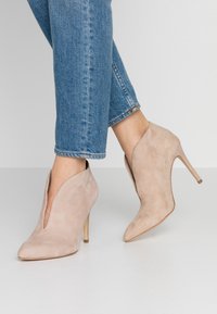 Béžové semišové kotníkové boty na vysokém podpatku s špičatou špičkou, otevřenými stranami a 10 cm stiletto podpatkem, v kombinaci s vyhrnutými modrými džíny.