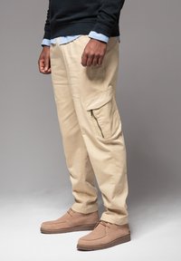 Beige Cargo-Hosen aus Baumwolle, mit einer Seitentasche, lockerer Passform und kombiniert mit braunen Wildlederschuhen. Strukturierter Stoff mit einer glatten Oberfläche.