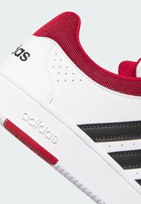 Białe buty sportowe z czerwonym siateczkowym kołnierzem, czarnymi paskami, teksturowanym boku i brandingiem. Gumowa podeszwa z czerwonymi akcentami i perforowanymi detalami.