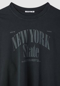 T-shirt nera grafica con scollo rotondo. Presenta la scritta "NEW YORK State" in grandi caratteri grigi e audaci, con ulteriori dettagli testuali sotto.