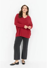 Crveni oversized V-izrez pulover s dugim rukavima, uparen s crnim skraćenim trapericama s frayed rubovima i crnim ravnim cipelama sjajnog završetka.