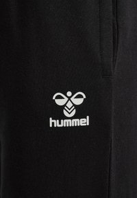 Schwarze Jogginghosen aus weichem Stoff mit einem weißen Logo und einem Emblem sowie dem Text "hummel" an der unteren Seite.