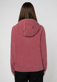 Person iført en støvet lyserød fleece-hoodie med grå trim, set bagfra, stående mod en ensfarvet lys baggrund.