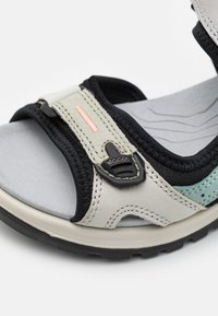 Sport sandal featuring a light gray leather upper, black textile straps with a pink accent, adjustable buckle, and cushioned gray footbed.
