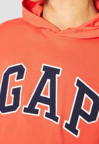 Primer plano de una persona vestida con una sudadera roja con grandes letras en azul marino y blanco que dicen "GAP" en el pecho.