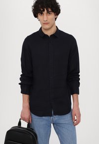 Donkerblauwe langemouwen shirt met knoopsluiting, zachte textuur en opgerolde mouwen. Het model houdt een zwarte rugzak vast en draagt lichtblauwe jeans.