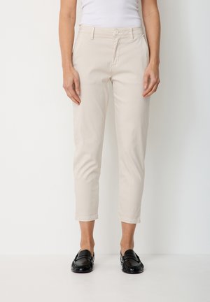 Frau trägt beige verkürzte Hose, schwarze Loafers und ein weißes ärmelloses Oberteil, steht vor einem einfachen weißen Hintergrund.