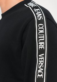 Melns džemperis ar ribotu tekstūru, kas iekļauj baltus drukātus akcentus pa piedurknēm ar uzrakstu "VERSACE JEANS COUTURE". Apaļa kakla izgriezne.