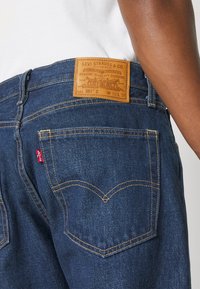 Gros plan d'un jean Levi's bleu foncé montrant la poche arrière, l'étiquette en cuir de la marque, ainsi qu'une partie du bras et de la chemise blanche d'une personne.
