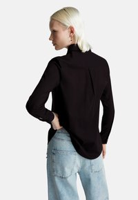 Donna con capelli biondi corti, che indossa una maglia nera a maniche lunghe e jeans azzurri chiari, in piedi con una mano sui fianchi, rivolta verso sinistra.