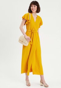 Yellow wrap dress with flutter sleeves, tied at the waist, featuring a deep V-neckline. Paired with a beige woven handbag and strappy heels.