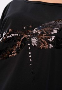 Zwarte shirt met een libelle ontwerp van pailletten in brons, zilver en zwart; soepele textuur met een wijde halslijn.