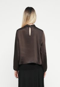 Woman with long blonde hair wearing a dark satin blouse with a back keyhole and button closure, and a black skirt, facing away.