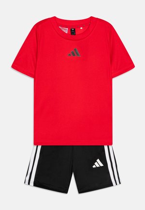 Rdeča kratkotrajna športna majica s reflektivnim črnim logotipom, v kombinaciji s črnimi kratkimi hlačami, ki imajo tri bele proge in ujemajoč logotip.